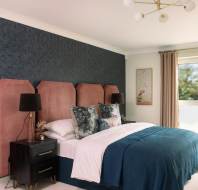 Bedroom with pink headboard, blue quilt, decorative pillows, black bedside tables, lamps, and a window with beige curtains.
