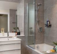Modern bathroom with gray tile walls, a glass shower door, a white sink with cabinet, a bathtub with showerhead, and accessories like soap and plants on the shelf.