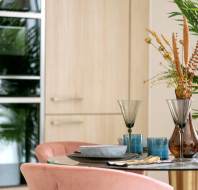 Dining table set with dishes and glasses, featuring a vase with dried flowers. Pink chairs surround the table. Wooden cabinets and green plants are in the background.