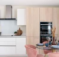 Modern kitchen with white cabinets, light wood paneling, and a stainless steel oven. There is a round dining table with pink chairs and tableware. A large green plant is in the corner.