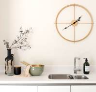Minimalist kitchen scene with white cabinets, a silver sink with a modern faucet, a beige wall clock, and decorative items including a black vase with dried branches and a green bowl.