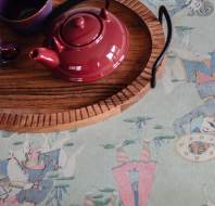A red teapot and a purple cup on a wooden tray, placed on a table with a colorful, artistic cloth featuring people in traditional attire.
