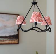 A black chandelier with three red-patterned shades hangs in a room with a painting on the wall and greenery below.