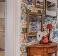 Wooden table with decorative items and floral arrangements stands against a wall with colorful wallpaper. Framed black and white photos hang above the table.