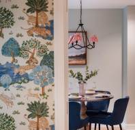 View through a doorway into a dining area with a round table, blue chairs, and a hanging light fixture. The wall on the left features colorful nature-themed wallpaper.