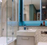 Modern bathroom with glass shower, illuminated mirror, wall-mounted sink, and toilet. Blue and beige color scheme with herringbone tile design and shelving.