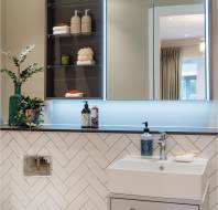 Modern bathroom with herringbone tiled wall, a white toilet, wall-mounted sink, illuminated mirror cabinet with toiletries, and a green plant. Doorway visible in the mirror reflection.