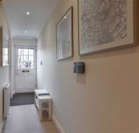 Narrow hallway with beige walls, framed maps, and a small bench. A closed white door with a window at the end lets in natural light.