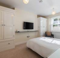 A bedroom with a large bed, two wardrobes, a wall-mounted TV, a chair by the window, and beige carpeting. Natural light enters through the window with blinds.