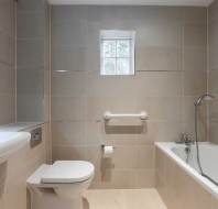 A beige-tiled bathroom features a wall-mounted sink with soap dispenser, a toilet, a bathtub with shower, and a small window above.