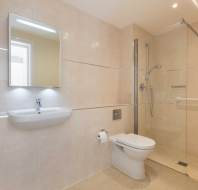 Modern bathroom with beige tiles, featuring a wall-mounted sink, mirror, toilet, and a glass-enclosed shower with a grab bar.