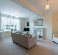 Living room with beige sofa, armchair, and a TV. A fireplace with a mirror above it is on the right, and a window with natural light brightens the space. A clock hangs on the wall.