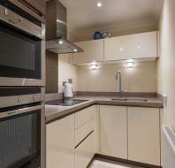 A compact modern kitchen with cream cabinets, an integrated oven, electric cooktop, and stainless steel sink. Bright lighting and minimal decoration create a clean look.