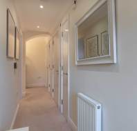 A narrow hallway with beige walls features doors on the left, framed pictures, and a radiator on the right. A large mirror hangs on the right wall.