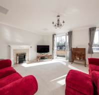 Living room with red sofas, fireplace, TV, chandelier, and large windows with gray curtains, allowing natural light. White walls and carpeted floor.