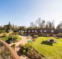 View from a balcony overlooking a garden with grass, a gazebo, a curved path, parked cars, a row of houses, and trees under a bright sky.