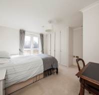 A clean, minimal bedroom with a double bed, gray accents, a desk with a chair, white wardrobe, and a window with curtains allowing natural light in.