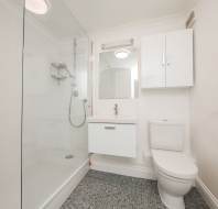 Modern bathroom with a glass-enclosed shower, white sink under a mirror, toilet, and tiled floor. White walls and cabinet create a clean, minimalist look.
