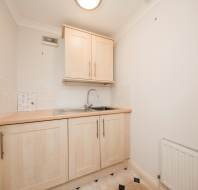 A small kitchen area with wooden cabinets, a stainless steel sink, beige countertop, white tiled backsplash, and a radiator below.