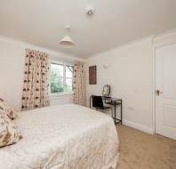 Bedroom with a double bed, floral bedding, and curtains. A window with a view of trees, a small desk with a chair, a decorative wall painting, and a white door are visible.