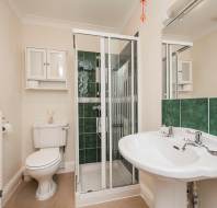 A bathroom with a shower cabin, white sink, toilet, and a wall-mounted cabinet. The walls are partially tiled in green, and a white towel rack is visible next to the toilet.