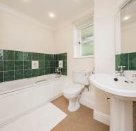 Bathroom with a white bathtub and green tiles, a toilet, and a white sink. A small window is above the toilet. The floor is covered with a beige carpet and a white mat.