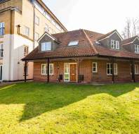 A brick house with a red tile roof and dormer windows is situated next to a multi-story apartment building. It is surrounded by a grassy lawn and trees in the background.