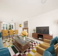 Spacious living room with a blue sofa, yellow armchairs, a large TV, and a dining table with yellow chairs. A colorful rug covers the floor, and a bouquet of flowers is on the glass coffee table.