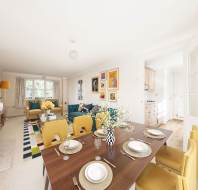 Bright, open-plan living and dining area with modern decor, featuring a table set for four, yellow chairs, a fireplace, colorful rug, and large windows. Adjacent kitchen visible through doorway.