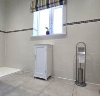 Modern bathroom with walk-in shower, white cabinet, floor-standing toilet paper holder, and a window with striped blinds. A small plant sits on the windowsill.