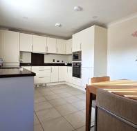 Modern kitchen with white cabinets, black countertops, and a built-in oven. A small dining table with chairs and two potted plants on the table and counter. Tile flooring and bright lighting.