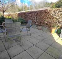 A stone-paved patio with four wicker chairs encircling a small table, surrounded by potted plants and a hedge. A leafless tree and blue sky are visible in the background.