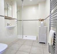 Modern bathroom with shower, white tiled walls, toilet, small sink with mirror, wicker cabinet with vase, and heated towel rail.