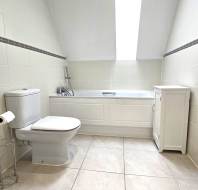Bathroom with white tiled walls, a skylight, toilet with a toilet paper holder, bathtub with a handheld showerhead, a small white cabinet, and beige floor tiles.
