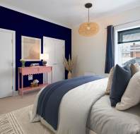 Modern bedroom with a blue accent wall, white bedding, and a beige headboard. A pink console table holds a lamp and artwork. A round wicker pendant light hangs from the ceiling.