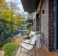 Modern balcony with glass railing, two chairs, a table, and potted plants. Trees with autumn foliage visible in the background.