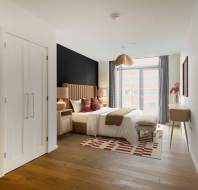 Modern bedroom with a large bed, side tables, and a bench on a geometric rug. Wooden floor, a window with curtains, and a pendant light overhead. A desk and chair are on the right.