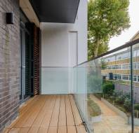Modern balcony with wooden decking and glass railing overlooking a tree-lined path and a brick building.