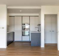 Modern, compact kitchen with gray cabinets, stainless steel appliances, and a sink. Wooden flooring extends into the adjoining room with an open door.