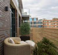 Balcony view with wicker seating, potted plants, and nearby apartment buildings under a cloudy sky.