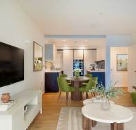 Modern open-plan living area with a TV, two round coffee tables, a green sofa, and green dining chairs. The kitchen, visible in the background, features dark cabinetry.