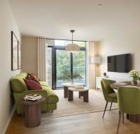 Modern living room with a green sofa, round dining table, and green chairs. Large window with curtains, wall-mounted TV, framed artworks, and a central light fixture. Wooden flooring and decor.