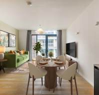 Modern open-concept living and dining area with a green sofa, wall art, and a TV. Table set for four, large window with curtains, and a potted plant. Light wooden flooring throughout.