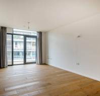 Empty room with wooden floors, white walls, and large window with curtains. Balcony view of building exterior.