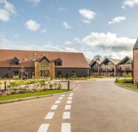 A modern residential complex with a main building in the center and additional structures to the right. The scene includes a road, landscaped gardens, and a partly cloudy sky.