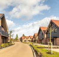 A suburban street with modern brick houses featuring sloped roofs and manicured lawns under a partly cloudy sky.