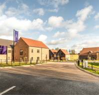 A suburban street scene shows several modern brick houses with pitched roofs, surrounded by lawns and fences, under a cloudy blue sky. Signs read "Shipston Meadows.