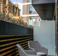 Two wicker chairs on a small outdoor balcony, with a frosted glass divider and a view of modern and traditional buildings under a partly cloudy sky.