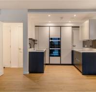Modern kitchen with wooden flooring, white walls, built-in appliances, and contrasting black and white cabinetry. Door on the left, small counters on either side. Bright overhead lighting.