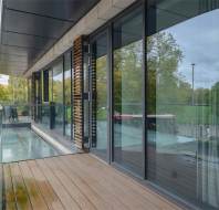 Modern balcony with wooden decking, clear glass railing, and large sliding glass doors reflecting trees and a bus in the background. A potted plant is at one end of the balcony.